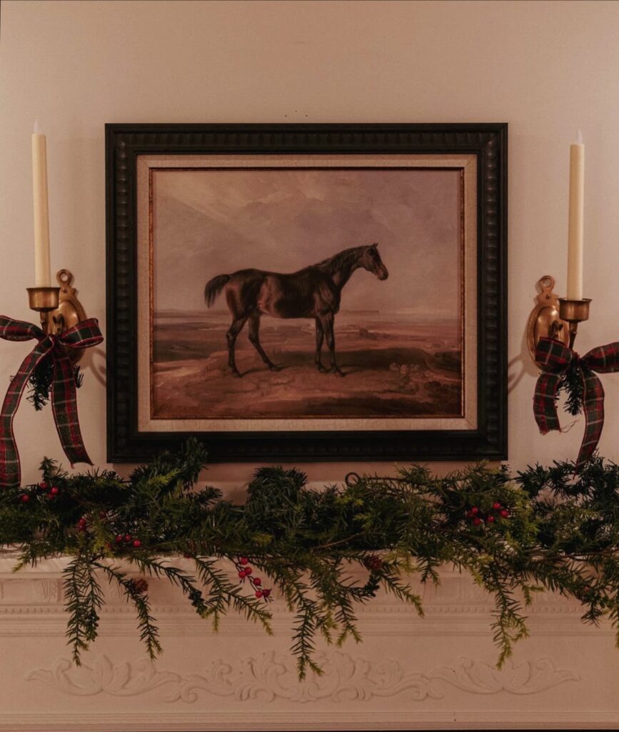 Evergreen garland with red berries and plaid bows decorating a white mantelpiece above a horse painting.