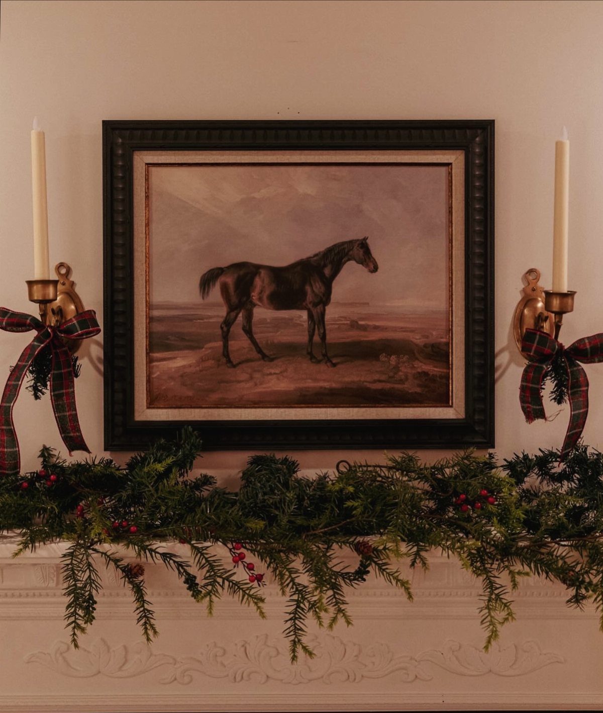 Evergreen garland with red berries and plaid bows decorating a white mantelpiece above a horse painting.
