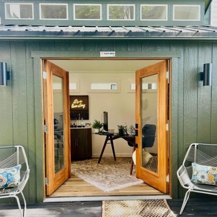 Green wooden shed with open glass doors revealing a modern home office interior.