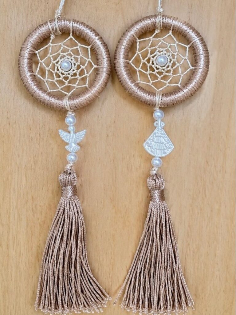 Two rose gold dreamcatcher ornaments with intricate webbing and long decorative tassels hanging on wood.