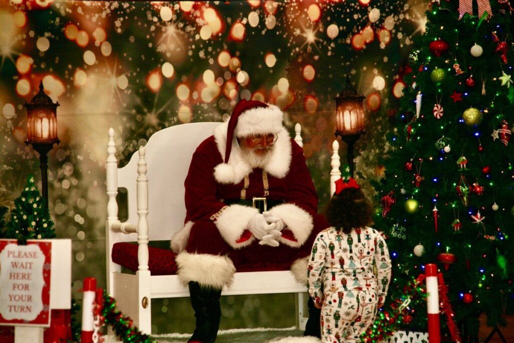 Santa Claus sitting in a white chair meeting a small child in festive pajamas.