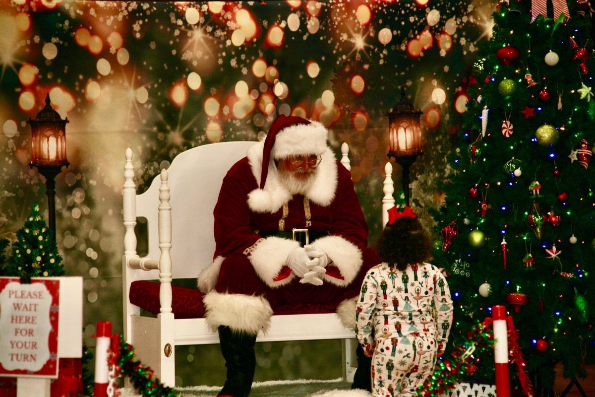 Santa Claus sitting in a white chair meeting a small child in festive pajamas.
