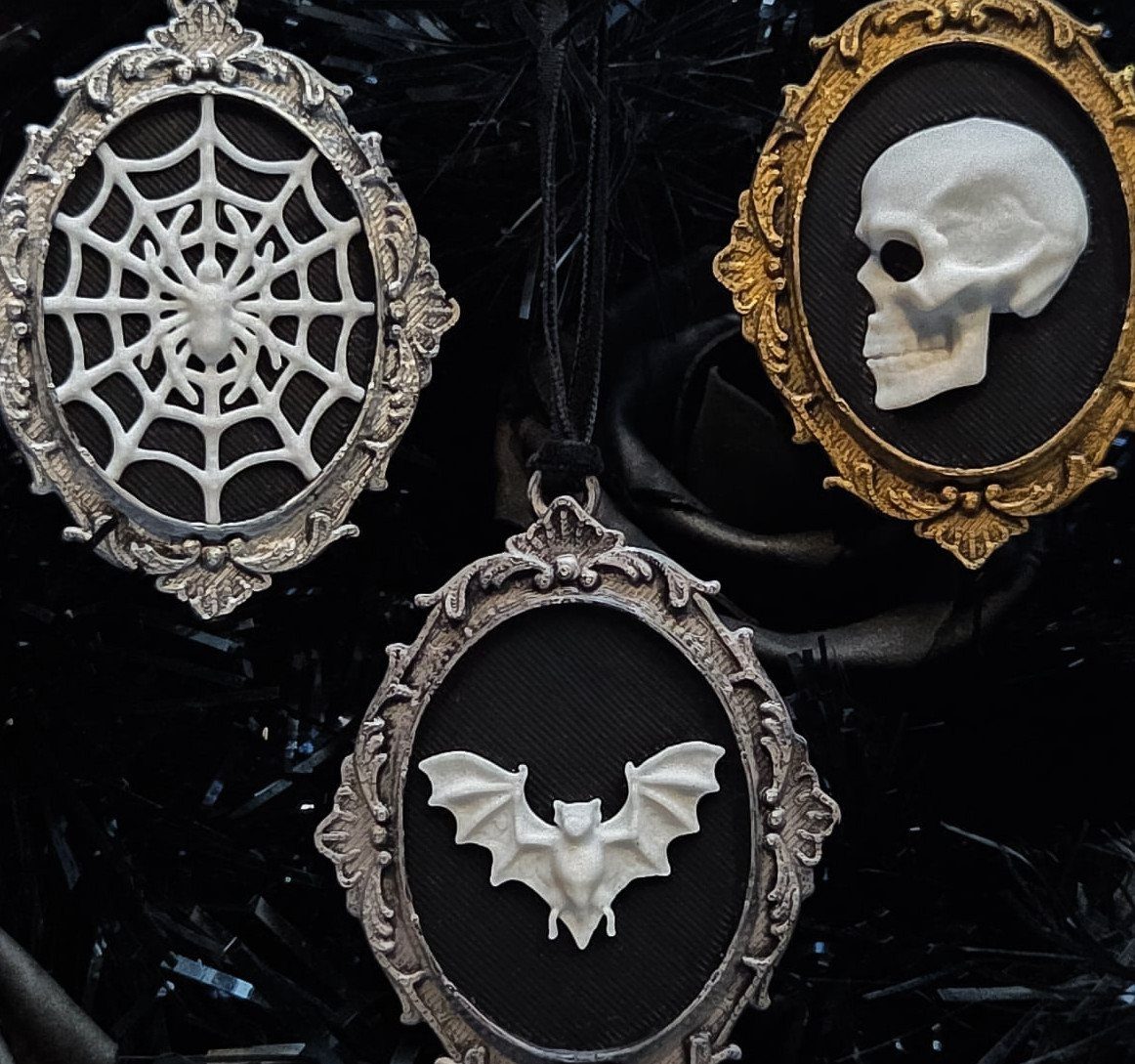Three ornate gothic ornaments featuring a spiderweb, skull, and bat hanging on a black tree.