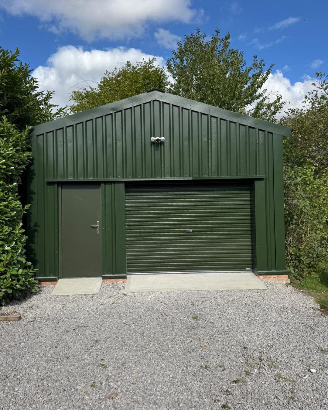 A large forest green metal garden building with a roller door and side door on gravel.